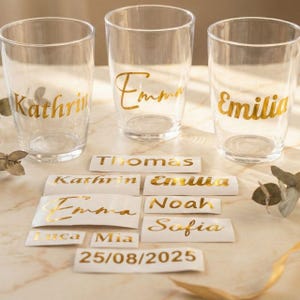 Könnte beinhalten: Drei transparente Gläser mit goldenen Buchstaben, die jeweils einen anderen Namen tragen: Kathrin, Emma und Emilia. Mehrere Namensaufkleber in goldener Schrift sind darunter angeordnet, zusammen mit einem Datum.