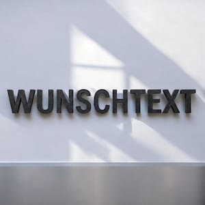 Könnte beinhalten: Dunkelgraue Buchstaben mit der Aufschrift "WUNSCHTEXT" sind an einer weißen Wand angebracht. Die Buchstaben werfen einen Schatten, und der Gesamteffekt ist modern und minimalistisch. Der Text ist ein deutsches Wort.