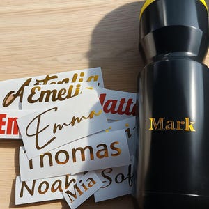 Könnte beinhalten: Eine schwarze Wasserflasche mit dem Namen "Mark" in goldfarbenen Buchstaben. Mehrere weiße Aufkleber mit goldenen Namen sind um die Flasche herum verstreut. Zu den Namen gehören Emma, Noah, Mia und andere. Die Flasche hat einen gelben Deckel.
