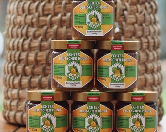 Puede incluir: Seis tarros de miel de vidrio con tapas doradas, etiquetados como "ECHTER DEUTSCHER HONIG" y "Sommertracht-honig". Cada tarro contiene 500g de miel, apilados frente a una cesta de mimbre.
