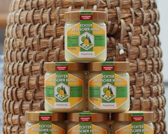 Puede incluir: Seis tarros de vidrio de miel etiquetados "Echter Deutscher Honig" con una etiqueta verde y amarilla. Los tarros están apilados en dos filas de tres. Cada tarro contiene 500 g de miel de colza.