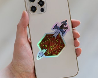 May include: A white iPhone with a holographic sticker of a 20-sided die with a sword piercing through it. The sticker has a rainbow iridescent finish.