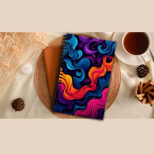 May include: A spiral-bound notebook with a vibrant abstract design in shades of blue, purple, orange, and pink. The notebook is placed on a woven tray with a brown notebook, a cup of tea, and colored pencils.