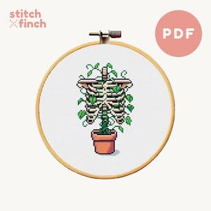 Puede incluir: Patrón de punto de cruz de una planta en maceta con una caja torácica esquelética que crece a través de ella. La planta tiene hojas verdes y está en una maceta marrón. El patrón está en un aro blanco.