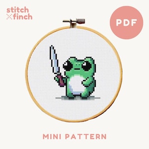 May include: A cross stitch pattern featuring a green frog holding a knife. The pattern is in a wooden embroidery hoop and the text "MINI PATTERN" is below the hoop. The text "PDF" is in a pink circle in the top right corner of the image.