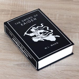 Puede incluir: Un libro de tapa dura titulado "THE SWORD OF KAIGEN" de M.L. Wang. El libro tiene una cubierta negra con texto blanco y una ilustraci&oacute;n blanca de una figura con una espada. El libro est&aacute; sobre una superficie de madera marr&oacute;n claro.