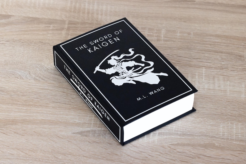 Puede incluir: Un libro de tapa dura titulado "THE SWORD OF KAIGEN" de M.L. Wang. El libro tiene una cubierta negra con texto blanco y una ilustraci&oacute;n blanca de una figura con una espada. El libro est&aacute; sobre una superficie de madera marr&oacute;n claro.