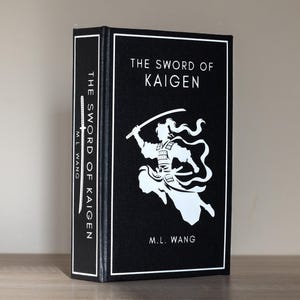 Puede incluir: Un libro de tapa dura titulado "The Sword of Kaigen" de M.L. Wang. El libro tiene una cubierta negra con texto blanco y una ilustraci&oacute;n blanca de un guerrero. El lomo tambi&eacute;n muestra el t&iacute;tulo y el autor.