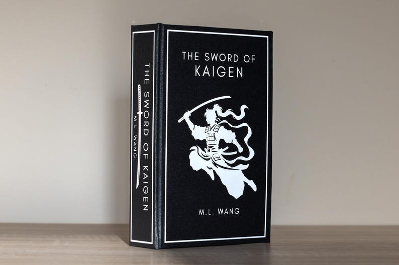 Puede incluir: Un libro de tapa dura titulado "The Sword of Kaigen" de M.L. Wang. El libro tiene una cubierta negra con texto blanco y una ilustraci&oacute;n blanca de un guerrero. El lomo tambi&eacute;n muestra el t&iacute;tulo y el autor.