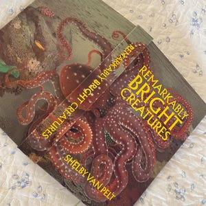 Op de afbeelding: Een hardcover boek getiteld Remarkably Bright Creatures door Shelby Van Pelt. De voorkant bevat een gedetailleerde illustratie van een grote, roodbruine octopus met witte stippen, geplaatst tegen een donkere, getextureerde achtergrond.