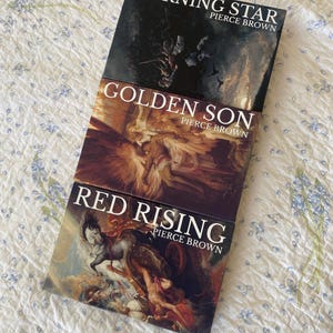May include: Three books by Pierce Brown stacked vertically. The top book is titled Morning Star, the middle book is Golden Son, and the bottom book is Red Rising. Each book cover features a detailed, classical-style painting with rich colors.