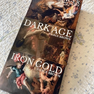 Pode incluir: Três livros de capa dura de Pierce Brown empilhados verticalmente. O livro superior intitula-se Light Bringer, o do meio Dark Age e o inferior Iron Gold. Cada capa apresenta uma pintura de estilo clássico com figuras em movimento.