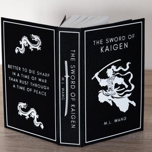 Puede incluir: Un libro de tapa dura titulado "The Sword of Kaigen" de M.L. Wang. El libro tiene una cubierta negra con texto e ilustraciones en blanco, incluyendo un drag&oacute;n y un guerrero. El lomo dice "The Sword of Kaigen".