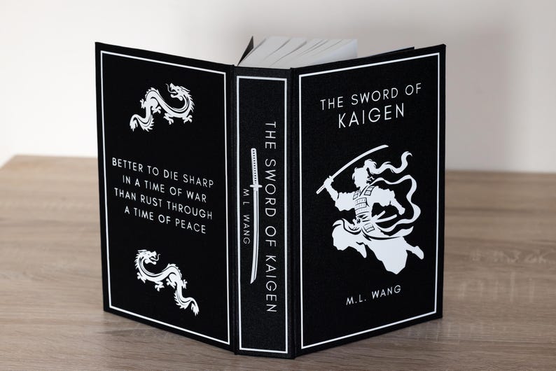 Puede incluir: Un libro de tapa dura titulado "The Sword of Kaigen" de M.L. Wang. El libro tiene una cubierta negra con texto e ilustraciones en blanco, incluyendo un drag&oacute;n y un guerrero. El lomo dice "The Sword of Kaigen".