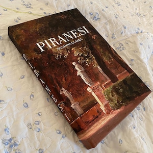 Puede incluir: Un libro de tapa dura titulado "PIRANESI" de Susanna Clarke. La portada presenta una pintura de una escena de jardín clásico con estatuas y vegetación. El libro está sobre una colcha con estampado floral blanco y azul.