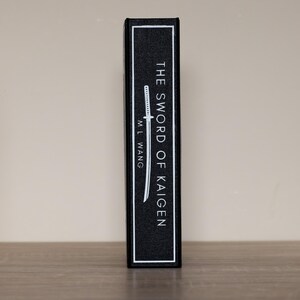 Puede incluir: Lomo de libro negro con texto blanco "THE SWORD OF KAIGEN" y el nombre del autor, "M.L. Wang". Tambi&eacute;n hay una ilustraci&oacute;n blanca de una espada. El libro est&aacute; sobre una superficie de madera.