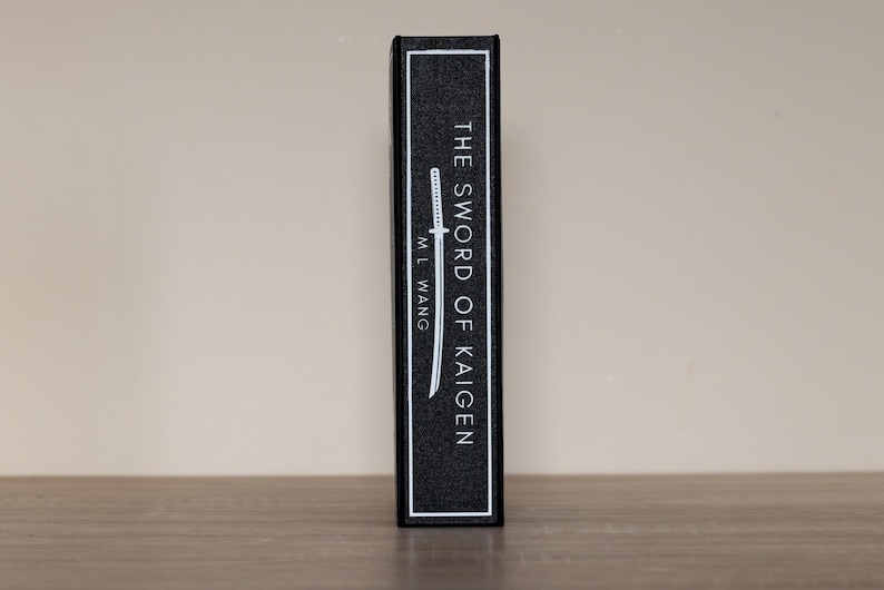 Puede incluir: Lomo de libro negro con texto blanco "THE SWORD OF KAIGEN" y el nombre del autor, "M.L. Wang". Tambi&eacute;n hay una ilustraci&oacute;n blanca de una espada. El libro est&aacute; sobre una superficie de madera.