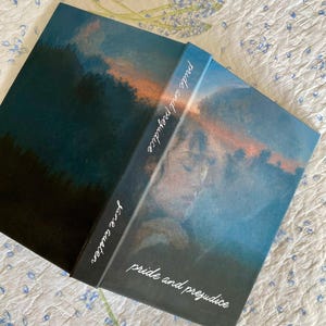 Peut inclure: Un livre de poche intitulé "Pride and Prejudice" de Jane Austen. La couverture présente une peinture de paysage dans les tons de bleu, orange et noir. Le titre et le nom de l'auteur sont en écriture blanche sur le dos et la couverture.