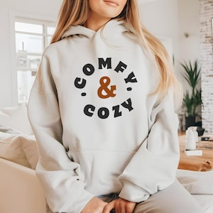 Könnte beinhalten: Ein cremefarbener Kapuzenpullover mit den Worten "COMFY & COZY" in einem kreisförmigen Design. Der Text ist in Schwarz und Braun gehalten, das kaufmännische Und in Braun. Der Hoodie hat eine Kapuze mit Kordelzug und lange Ärmel.