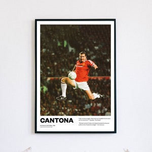 May include: A framed poster showing a football player in a red jersey kicking a ball. The poster has the name "CANTONA" in bold white letters. The image is set against a white wall. The poster includes a quote.