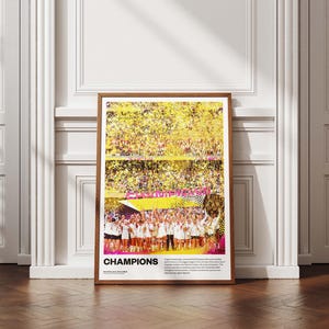 May include: A framed poster with a wooden frame, featuring a vibrant image of a sports team celebrating a victory. The word "CHAMPIONS" is prominently displayed in pink and white. The background shows a stadium filled with confetti.