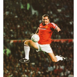 May include: A vintage-style poster of Eric Cantona in a Manchester United kit, mid-air with a football. The poster features the name "CANTONA" and quotes by Roy Keane and Eric Cantona. The image has a retro aesthetic, with a blurred background.