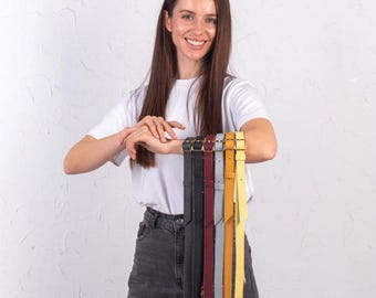 May include: A woman is holding up a variety of leather belts in different colors, including black, brown, gray, yellow, and red. The belts are all made of leather and have a simple design.