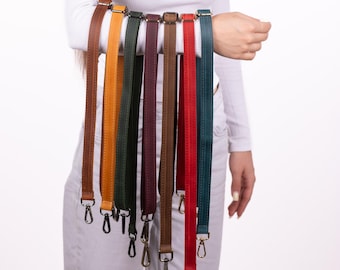 Op de afbeelding: Een collectie van zeven lange, platte leren tashengsels in diverse kleuren, waaronder bruin, geel, donkergroen, bordeauxrood, beige, rood en blauwgroen. Elke riem is voorzien van gestikte randen en metalen draaibare karabijnsluitingen.