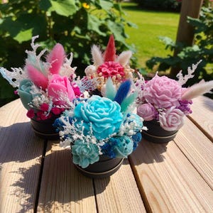 Può includere: Quattro piccoli contenitori neri riempiti con composizioni floreali decorative e colorate. Le composizioni presentano fiori di sapone scolpiti nelle tonalità del rosa, del blu e del viola, accentuati da fiori secchi e piume. I contenitori sono su una superficie di legno.