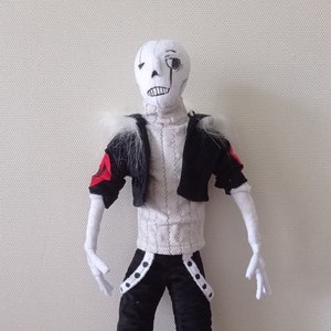 Puede incluir: Una muñeca de peluche blanca y negra con una chaqueta roja y negra, un suéter blanco, pantalones negros y botas grises. La muñeca tiene una cara blanca con ojos negros y un parche negro en el ojo.