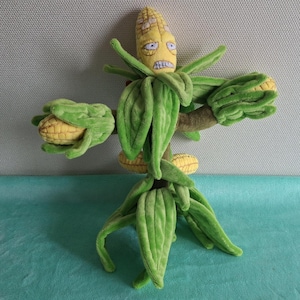 Op de afbeelding: Een pluche maïskarakter met een geel gezicht, groene bladeren en armen die maïskolven vasthouden. Het personage heeft een boze uitdrukking en staat rechtop. Het pluche speelgoed staat tegen een lichtgrijze en blauwgroene achtergrond.