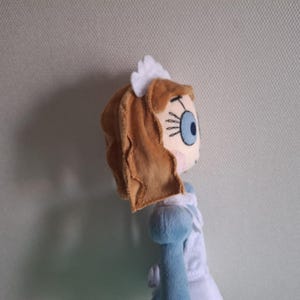 May include: A plush doll featuring a character with light brown hair, a blue eye, and a white headpiece. The doll is wearing a blue and white dress. The doll's face is detailed with stitched features.
