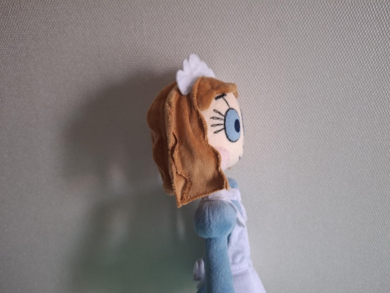 May include: A plush doll featuring a character with light brown hair, a blue eye, and a white headpiece. The doll is wearing a blue and white dress. The doll's face is detailed with stitched features.