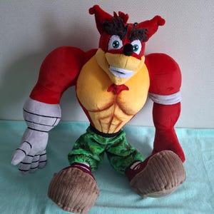 Può includere: Un peluche di un personaggio dei cartoni animati con pelliccia rossa, petto giallo e pantaloni mimetici verdi. Il personaggio ha un ampio sorriso, sopracciglia marroni e grandi piedi marroni. Il giocattolo è alto circa 50 cm.