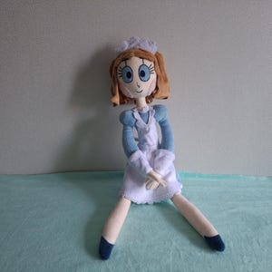 May include: A handmade doll with a light skin tone, wearing a blue and white dress, a white apron, and a white cap. The doll has brown hair, blue eyes, and is sitting with its legs spread. The doll's shoes are dark blue.