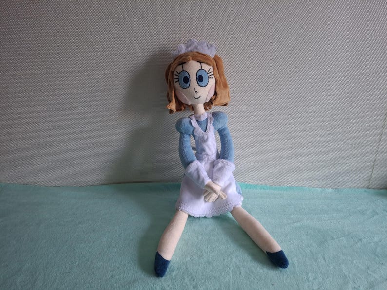 May include: A handmade doll with a light skin tone, wearing a blue and white dress, a white apron, and a white cap. The doll has brown hair, blue eyes, and is sitting with its legs spread. The doll's shoes are dark blue.