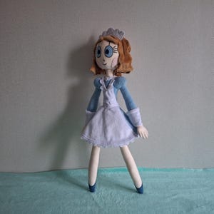 May include: A handmade doll with a blue and white dress, blue shoes, and brown hair. The doll has a white apron, blue sleeves, and a small crown. The doll's face has large blue eyes and rosy cheeks. The doll is standing on a light blue surface.
