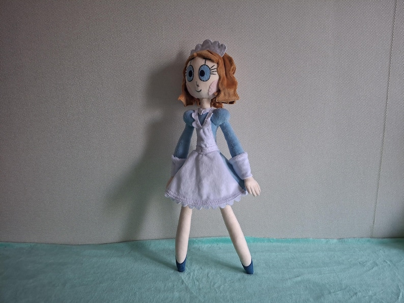 May include: A handmade doll with a blue and white dress, blue shoes, and brown hair. The doll has a white apron, blue sleeves, and a small crown. The doll's face has large blue eyes and rosy cheeks. The doll is standing on a light blue surface.