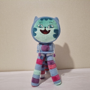 Puede incluir: Un peluche de gato con cabeza azul y cara sonriente, sobre una base de trípode con patas a rayas en tonos azules, rosas y morados. La cabeza del gato tiene una capucha azul claro y detalles turquesa.