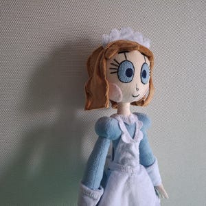 May include: A handmade doll with a light brown hairstyle, large blue eyes, and rosy cheeks. The doll wears a light blue dress with a white apron and a white headpiece. The doll is a soft, plush toy.
