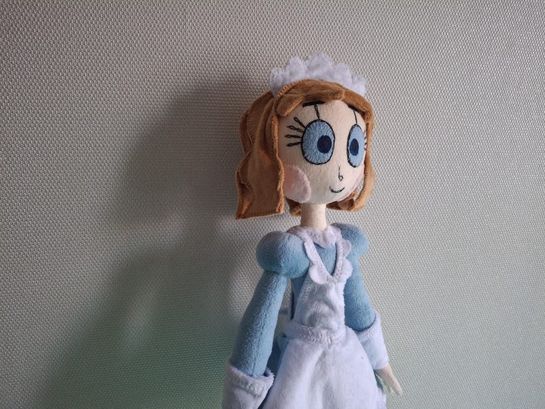 May include: A handmade doll with a light brown hairstyle, large blue eyes, and rosy cheeks. The doll wears a light blue dress with a white apron and a white headpiece. The doll is a soft, plush toy.