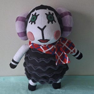 Può includere: Un giocattolo di peluche di pecora nero e bianco con orecchie viola, una sciarpa a quadri rossi e neri e grandi occhi verdi. La pecora indossa un vestito nero con un orlo arricciato.