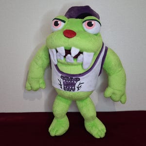 Puede incluir: Juguete de peluche verde con un sombrero morado y un tanque blanco con un diseño morado. El juguete tiene una boca grande con dientes blancos y una nariz roja.