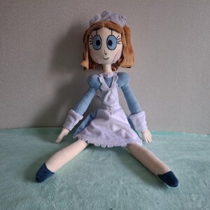 May include: A handmade doll dressed in a light blue and white maid outfit. The doll has brown hair, blue eyes, and a white cap. The doll's legs are spread apart, and it is sitting on a light blue surface.