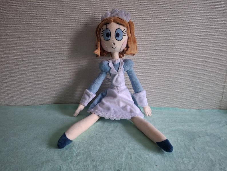 May include: A handmade doll dressed in a light blue and white maid outfit. The doll has brown hair, blue eyes, and a white cap. The doll's legs are spread apart, and it is sitting on a light blue surface.