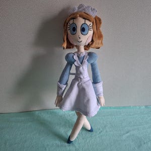 May include: A handmade plush doll featuring a cartoon-style design. The doll has light brown hair, large blue eyes, and is dressed in a light blue and white outfit with a matching cap. The doll is standing on a light blue surface.
