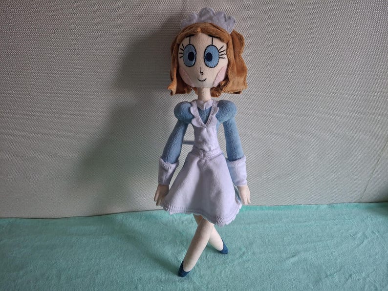 May include: A handmade plush doll featuring a cartoon-style design. The doll has light brown hair, large blue eyes, and is dressed in a light blue and white outfit with a matching cap. The doll is standing on a light blue surface.