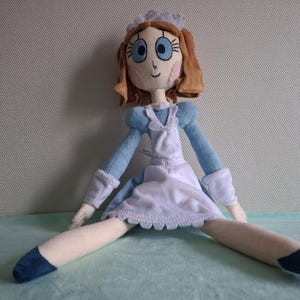 May include: A handmade doll with a light blue dress, white apron, and a matching bonnet. The doll has brown hair, blue eyes, and is sitting with its legs spread. The doll's shoes are dark blue.
