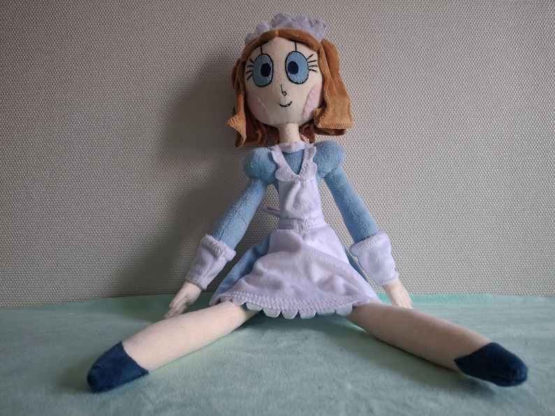 May include: A handmade doll with a light blue dress, white apron, and a matching bonnet. The doll has brown hair, blue eyes, and is sitting with its legs spread. The doll's shoes are dark blue.