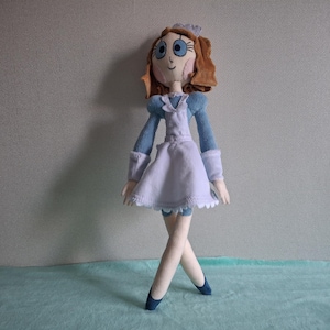 May include: A handmade doll in a maid outfit. The doll has brown hair, blue eyes, and a light skin tone. The outfit is light blue and white, with a white apron and a matching bonnet. The doll is standing with its legs crossed and wearing blue shoes.
